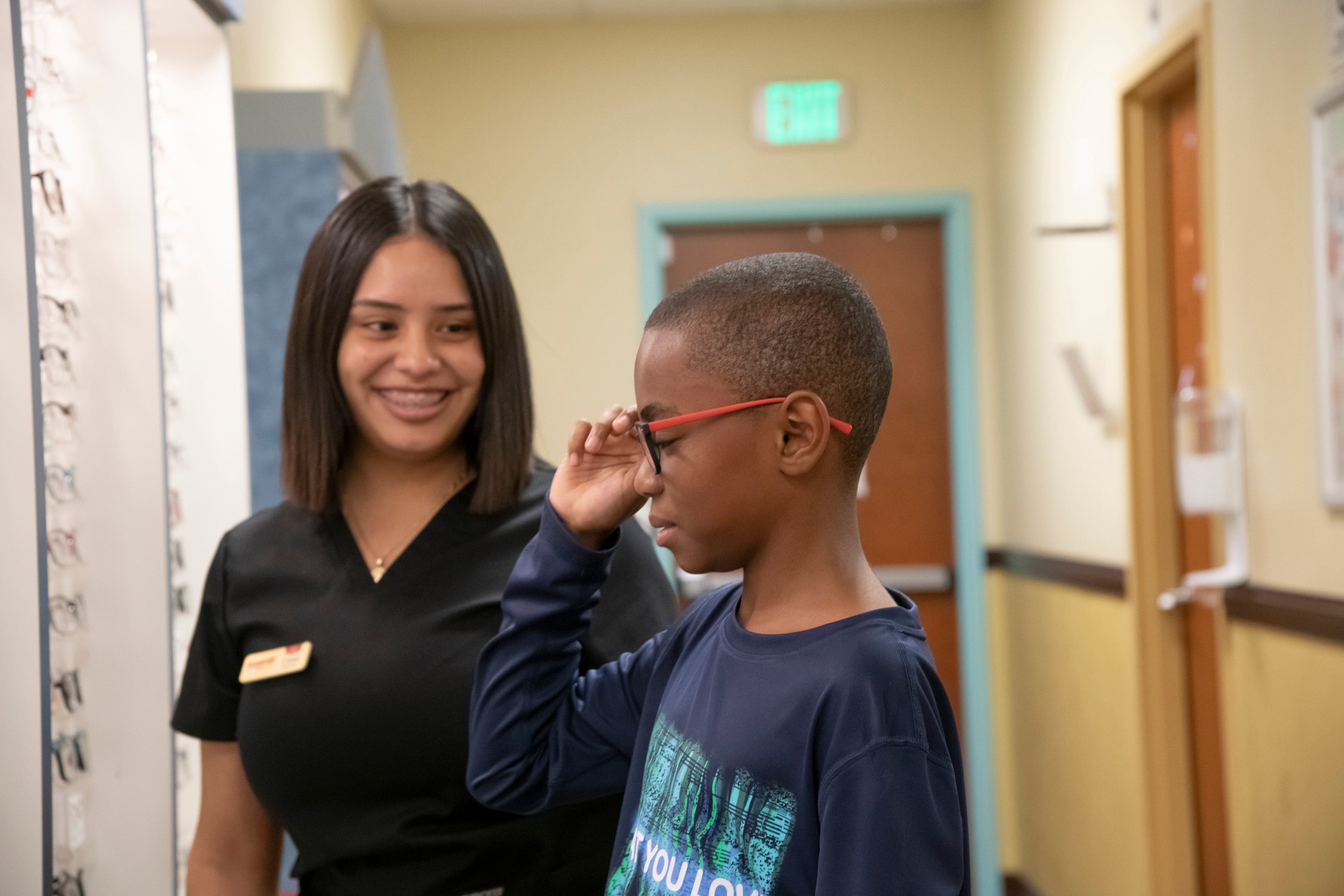 young boy wearing a blue shirt trying on red children's glasses with help from staff member