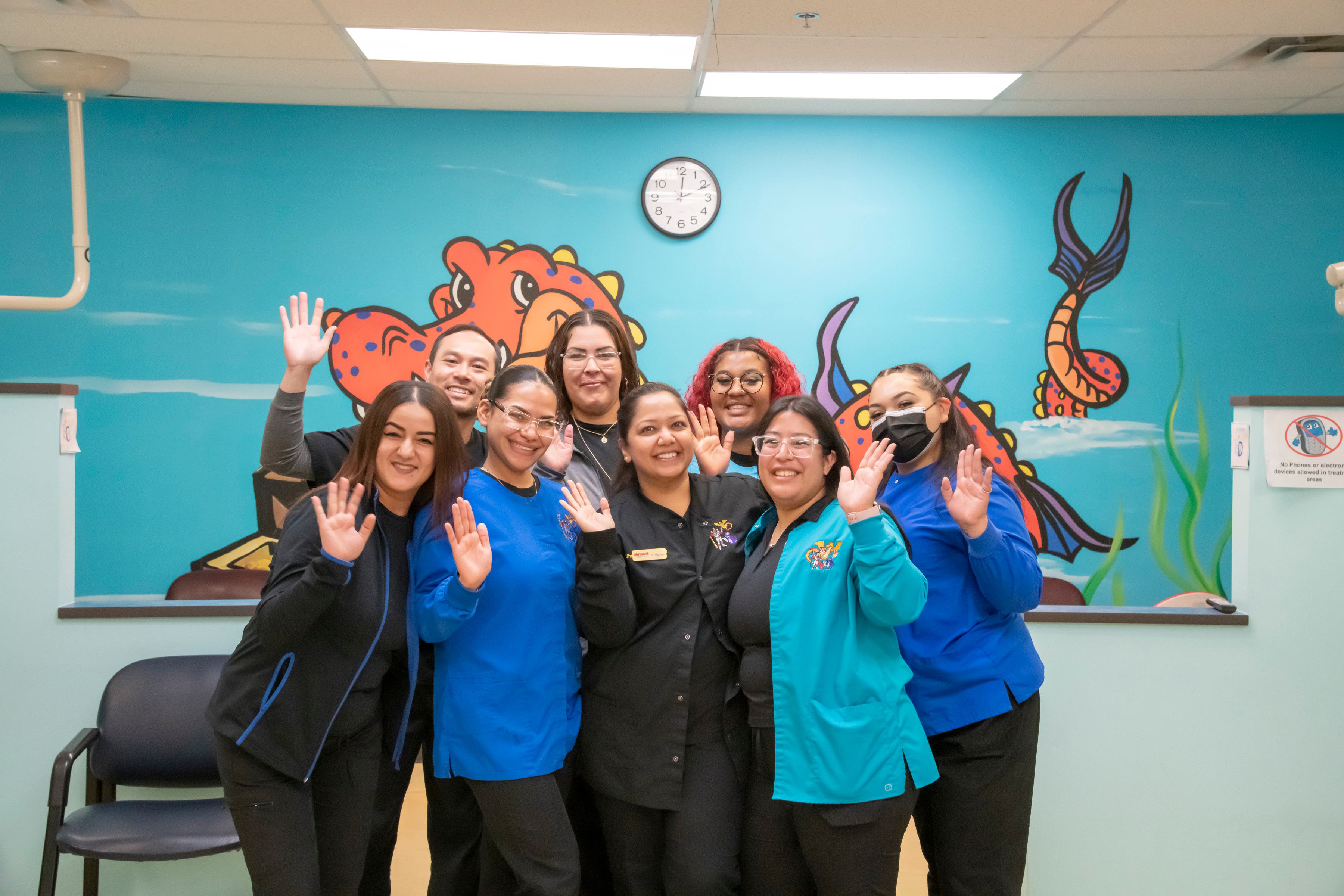 Pediatric dental and vision care staff at our east colfax location in Aurora. There are 8 people in the photo and they're waving and wearing blue clothing.