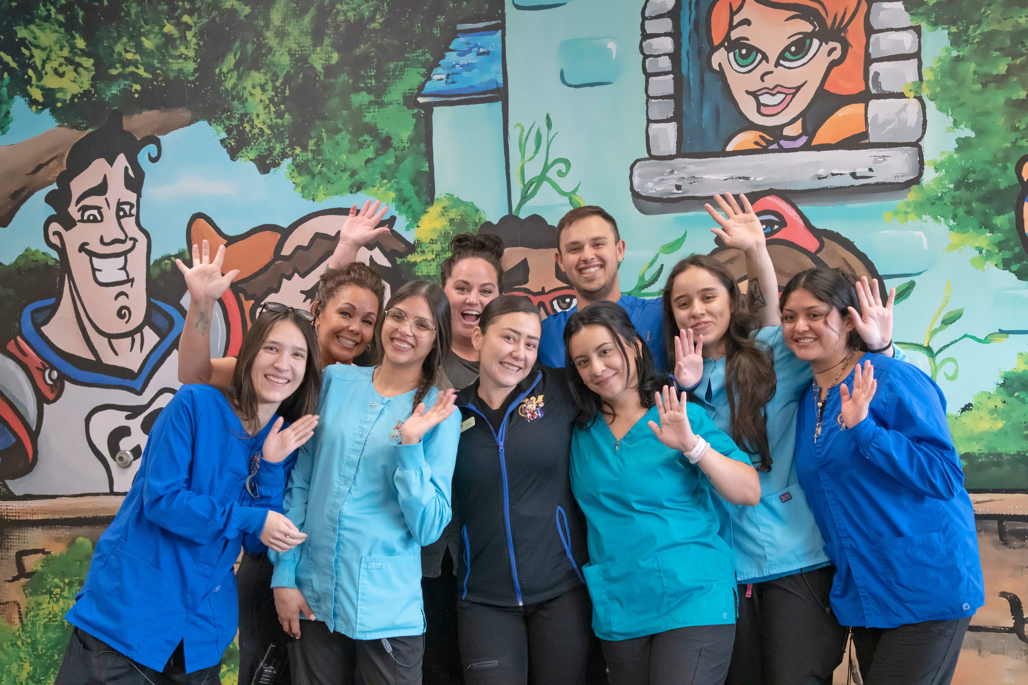 Nine staff members of our pediatric dental, vision and orthodontics team are waving in front of a colorful background. They are at our Lakwood location.