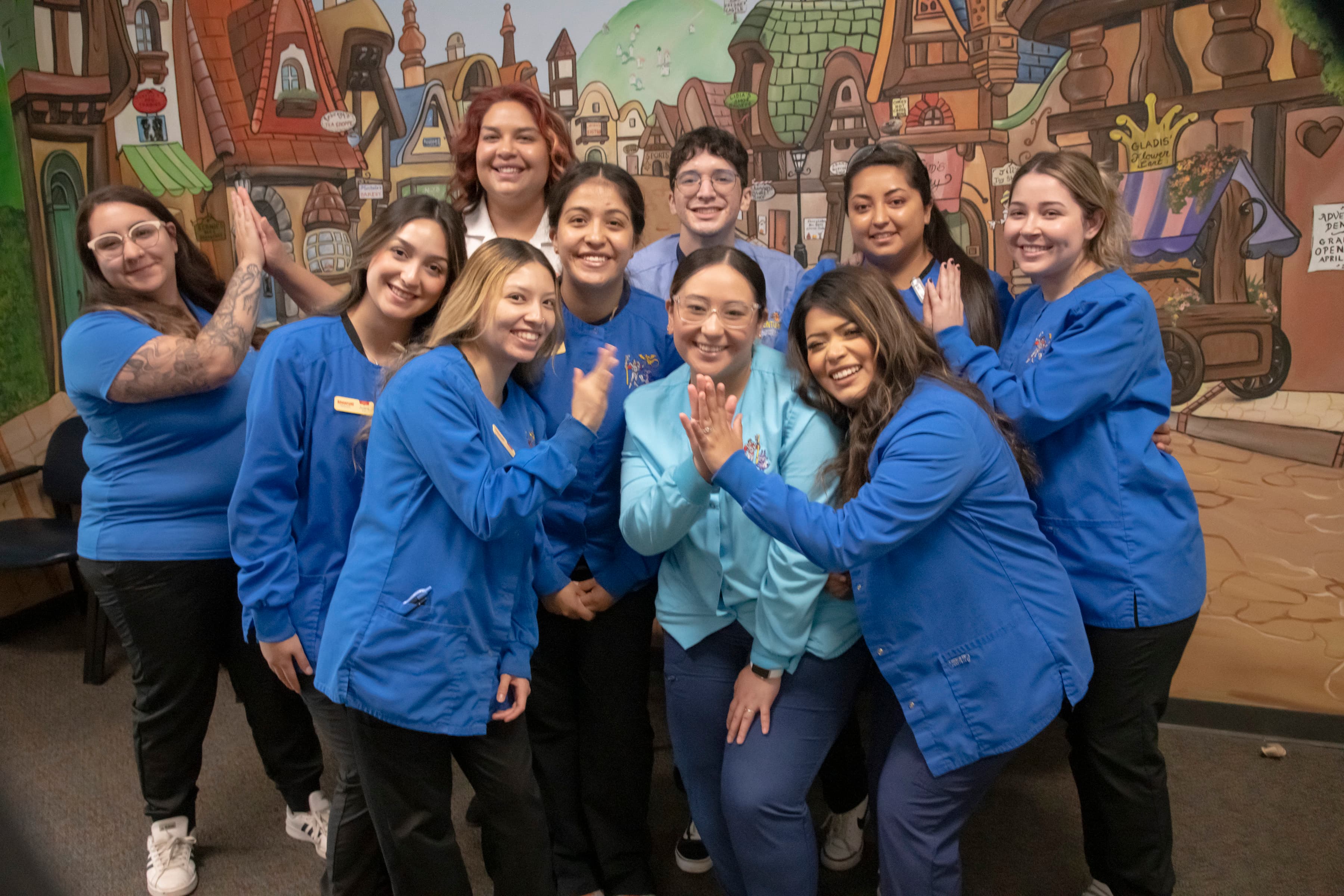 Pediatric dentistry, visions and orthodontic healthcare professionals at our Longmont office. Nine woman are waving and smiling wearing blue uniforms.