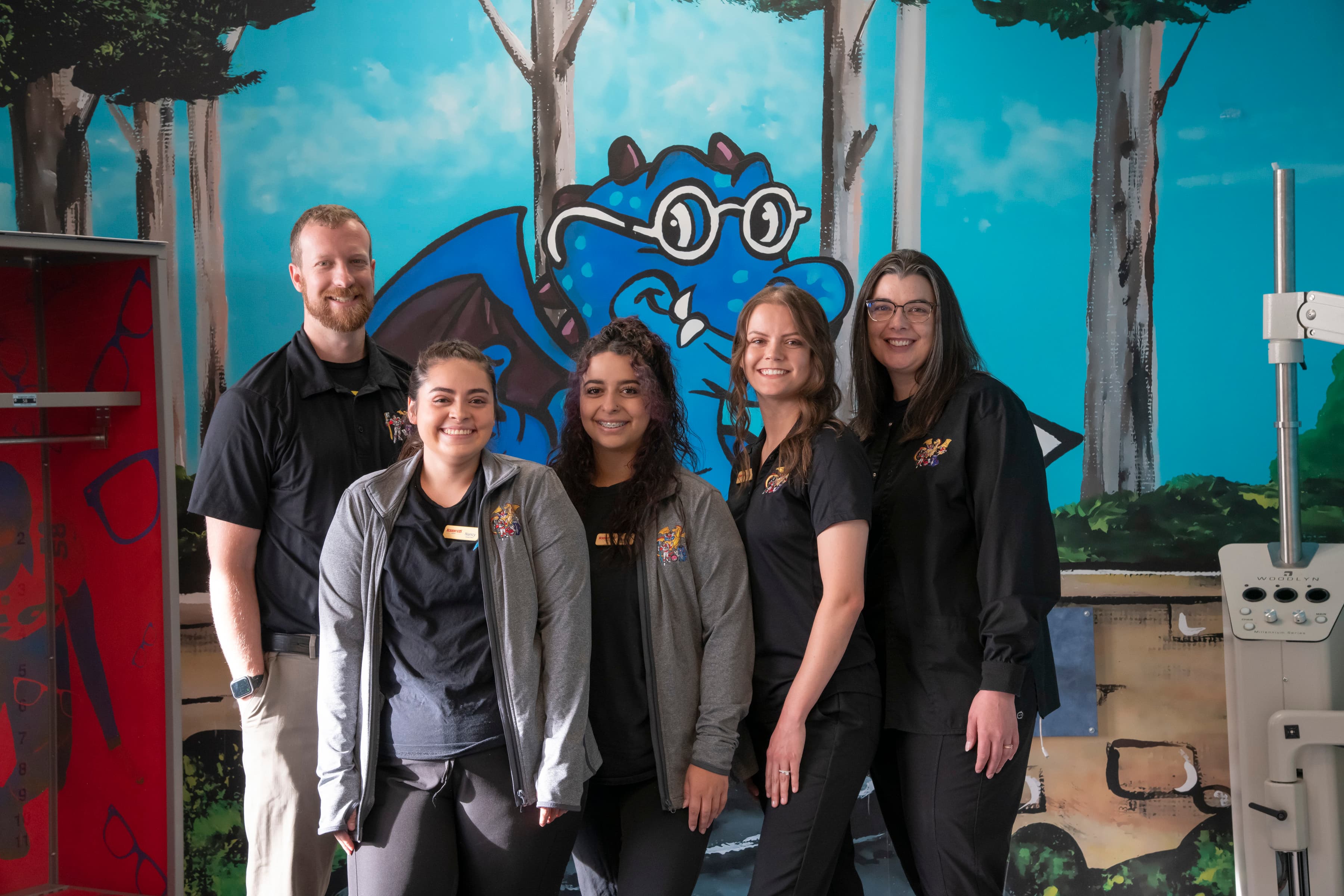 Kid's Optometrist staff at our Pueblo location. There are five people in wearing black and grey medical attire and are standing in front of a kids cartoon.